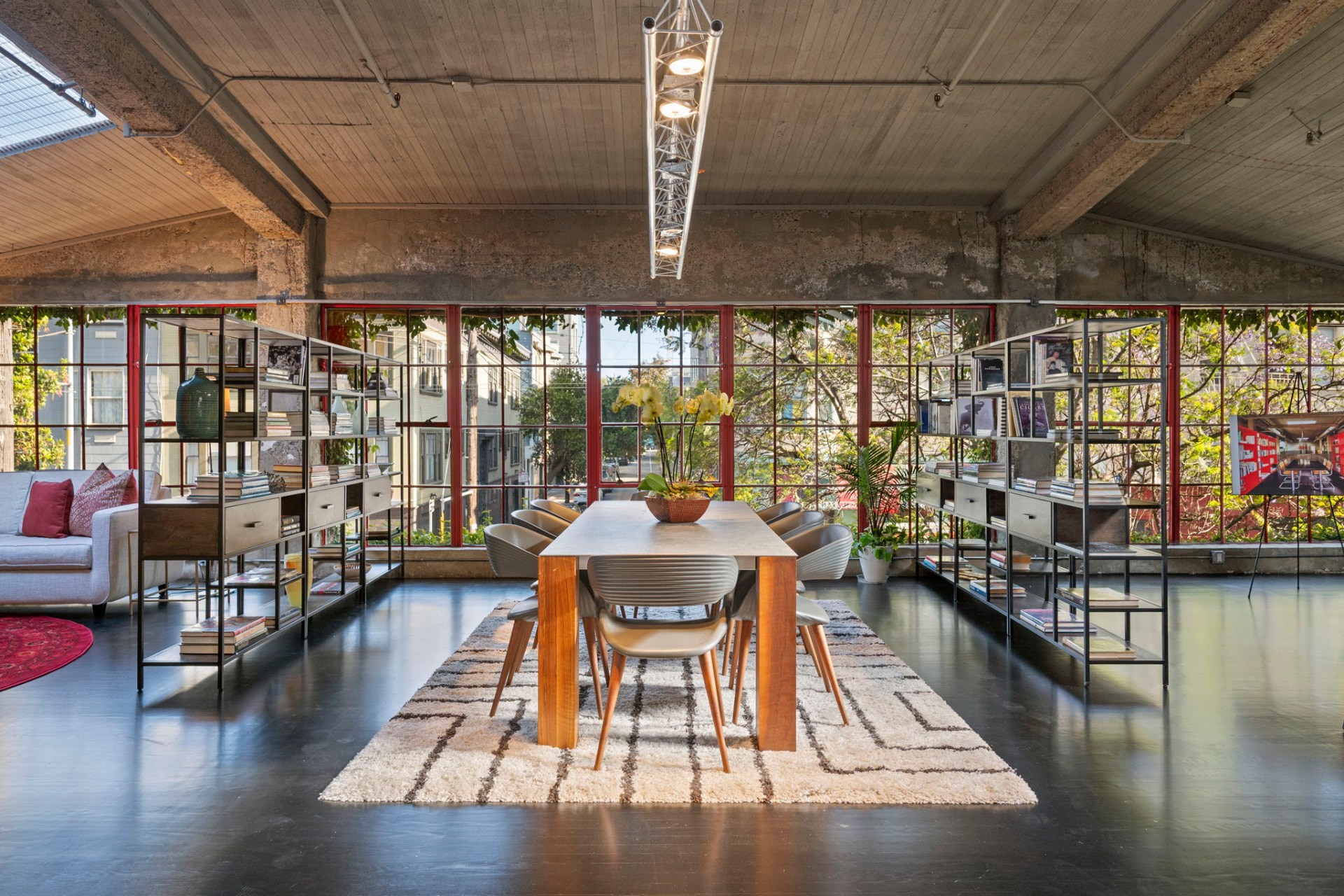 Long communal table beneath a truss light fixture, flanked by steel bookshelves and a wall of red-framed factory windows