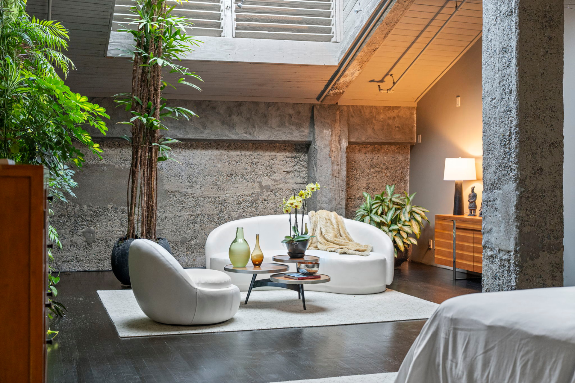 Quiet sitting area with a curved white sofa against a raw concrete wall, tall plants and a skylight above